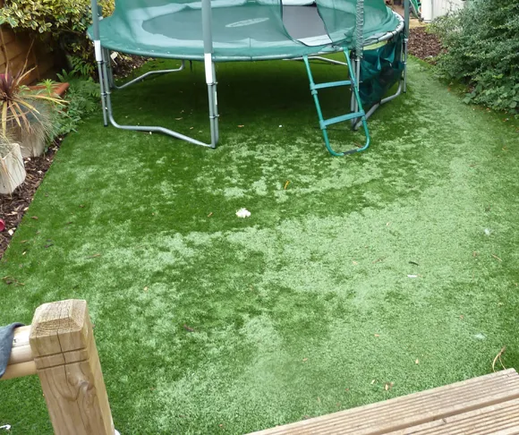 Residential garden with worn artificial grass and a large trampoline, showing signs of weathering and usage, surrounded by lush greenery and a wooden fence.
