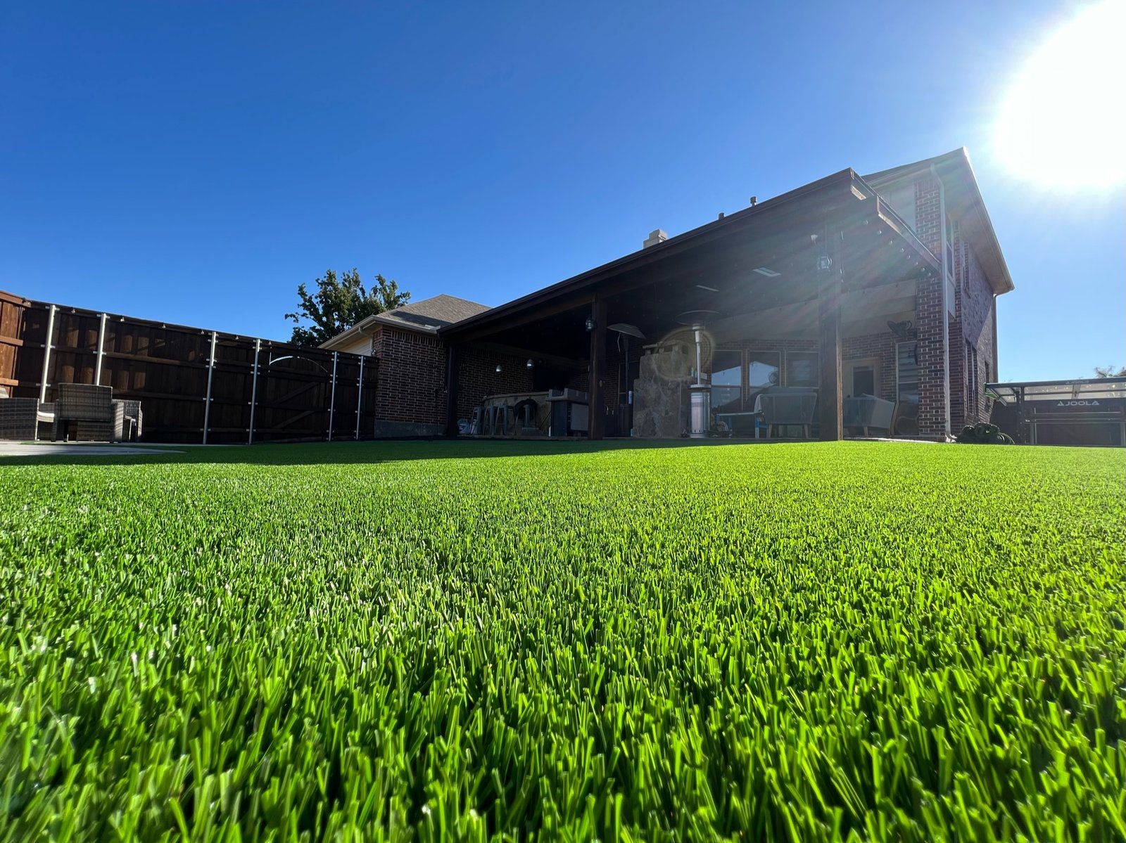 Vibrant artificial grass in the foreground with a residential home and covered patio in the background, under a clear blue sky, showcasing a luxurious home exterior and landscaping.