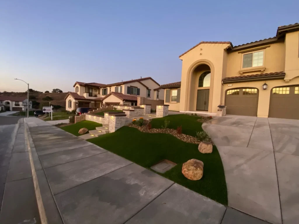 Elegant suburban homes with manicured landscaping, synthetic lawn, and decorative stonework along a paved driveway, captured at twilight.