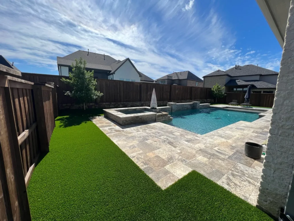Inviting backyard with a swimming pool and fountain feature and patio pavers, bordered by a wooden fence and artificial grass, under a blue sky with fluffy clouds.