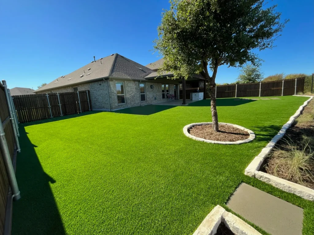 Well-maintained backyard with vibrant green artificial turf and a mature tree in a mulched bed, edged by curved stone borders, adjacent to a stone residential home.