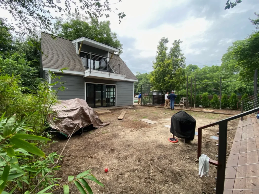 Backyard in early stages of landscaping with bare soil, a modern grey home with upper balcony, covered outdoor items, and two people working near a metal fence.