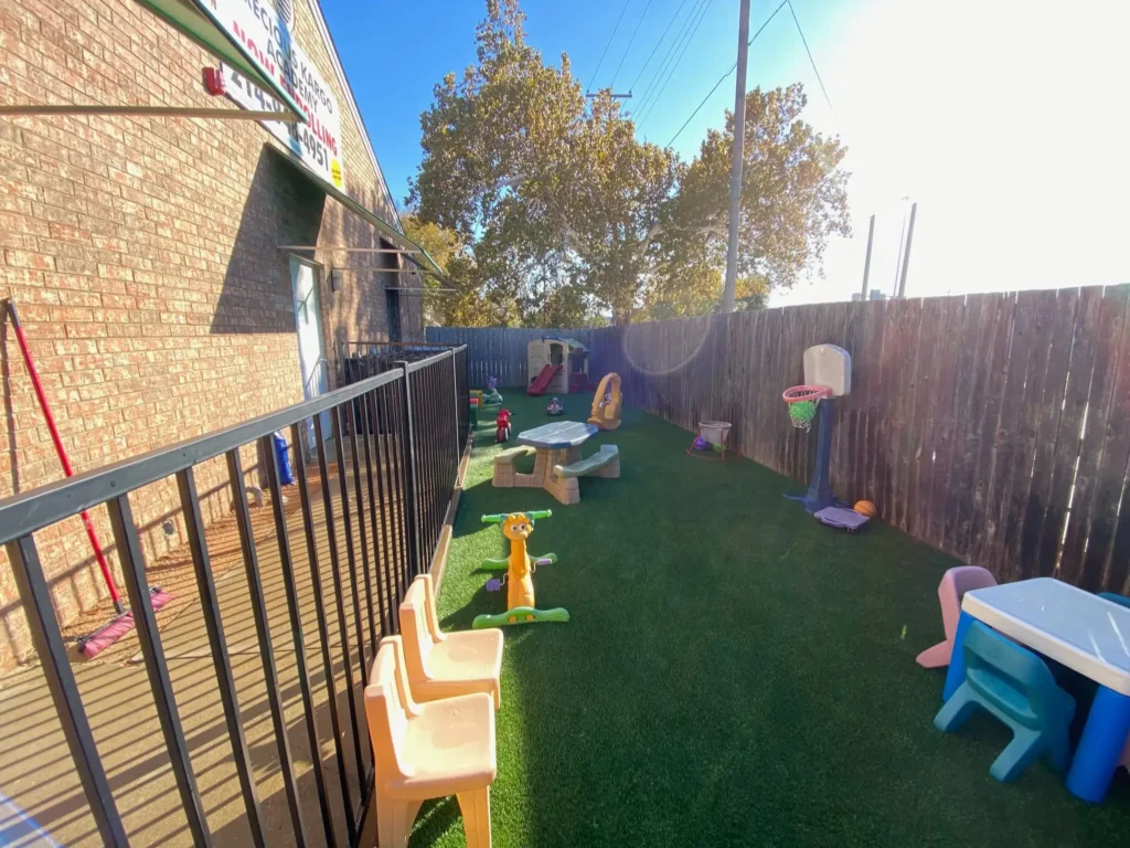 Children's play area in a daycare with artificial grass and various toys, including slides, play tables, and basketball hoops, enclosed by a safety fence and a brick building.