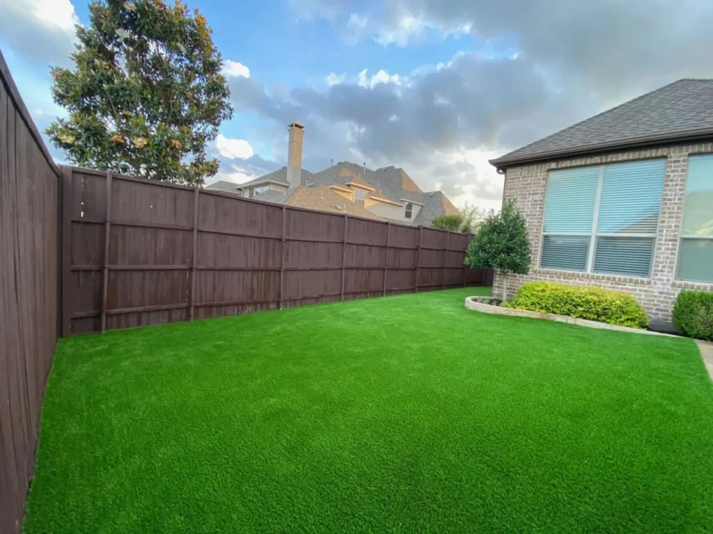 Spacious backyard with freshly installed artificial turf, bordered by a tall wooden fence, with a residential brick house and landscaped garden bed under a cloudy sky.