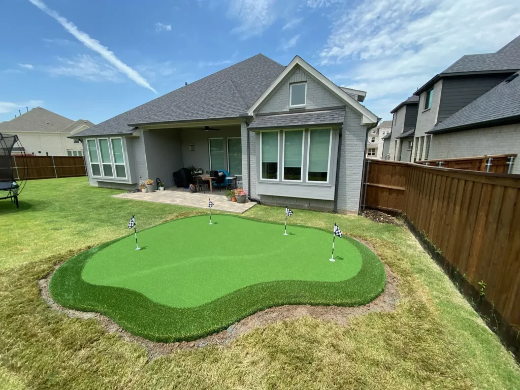 Spacious residential backyard with a custom artificial grass putting green, checkered flagsticks, surrounded by natural grass and a wooden fence, with a view of the home's gray exterior and patio.