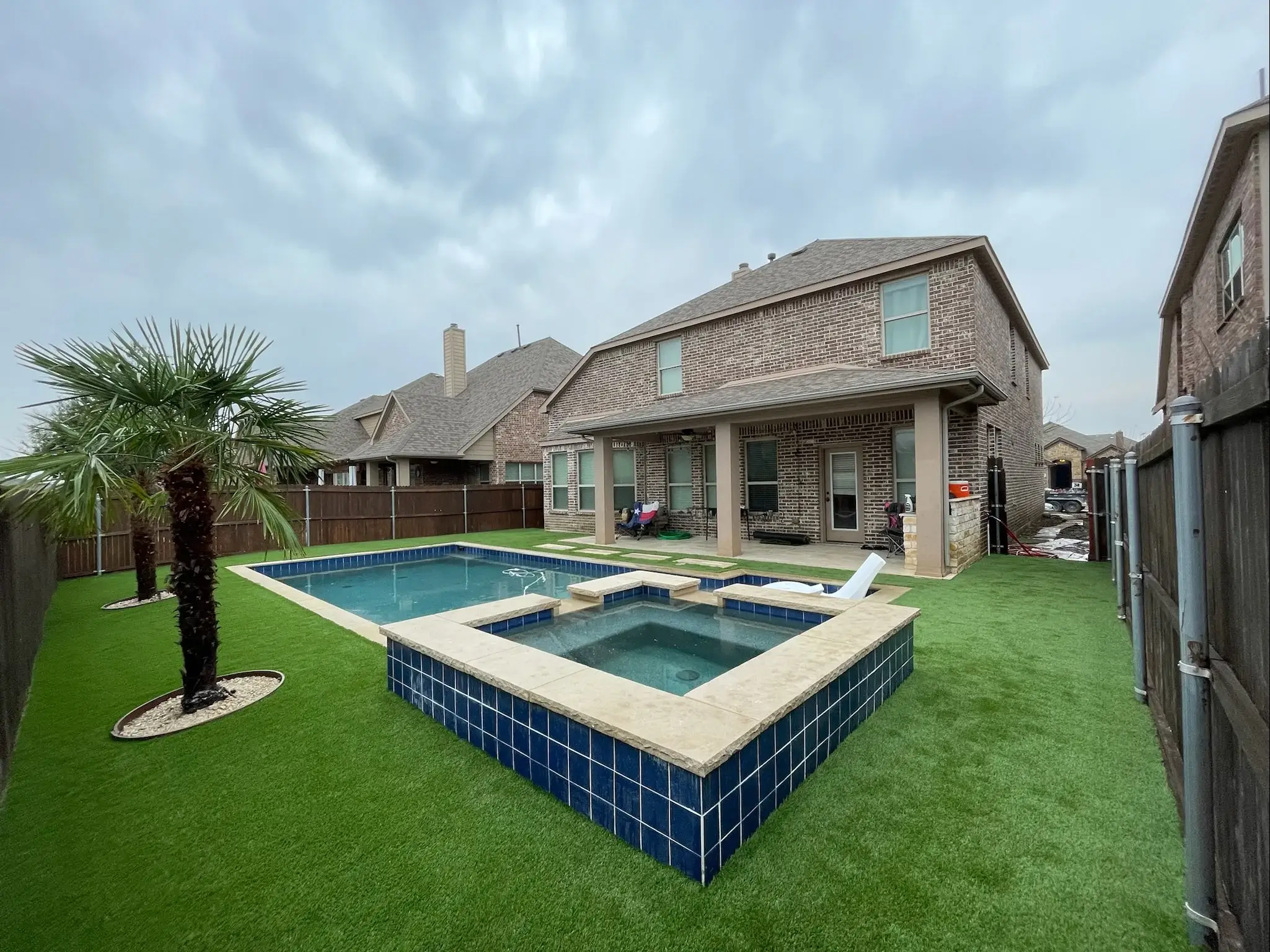 A backyard scene of a suburban home featuring a well-maintained, artificial turf lawn. In the center is a rectangular swimming pool lined with blue tiles and surrounded by a stone deck. Two palm trees enhance the landscape, suggesting a comfortable, leisurely outdoor space. The overcast sky above and a glimpse of the neighborhood beyond the wooden fence suggest a tranquil, residential setting.