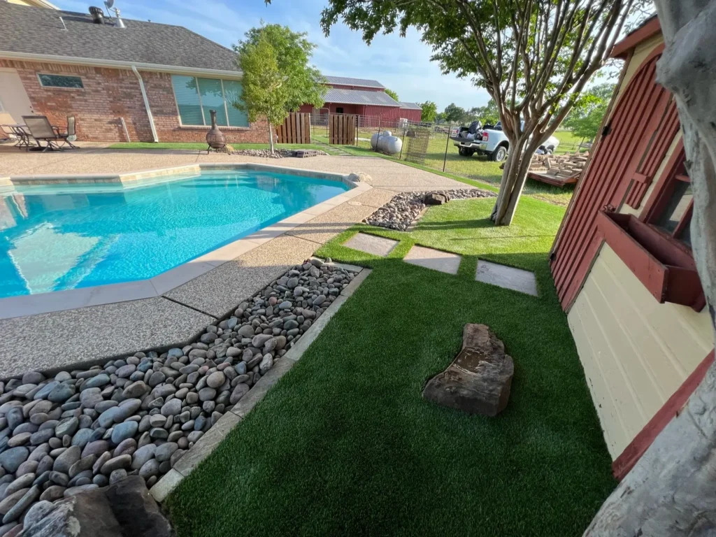 Well-manicured backyard with a swimming pool, artificial turf, and decorative rocks under a clear blue sky.