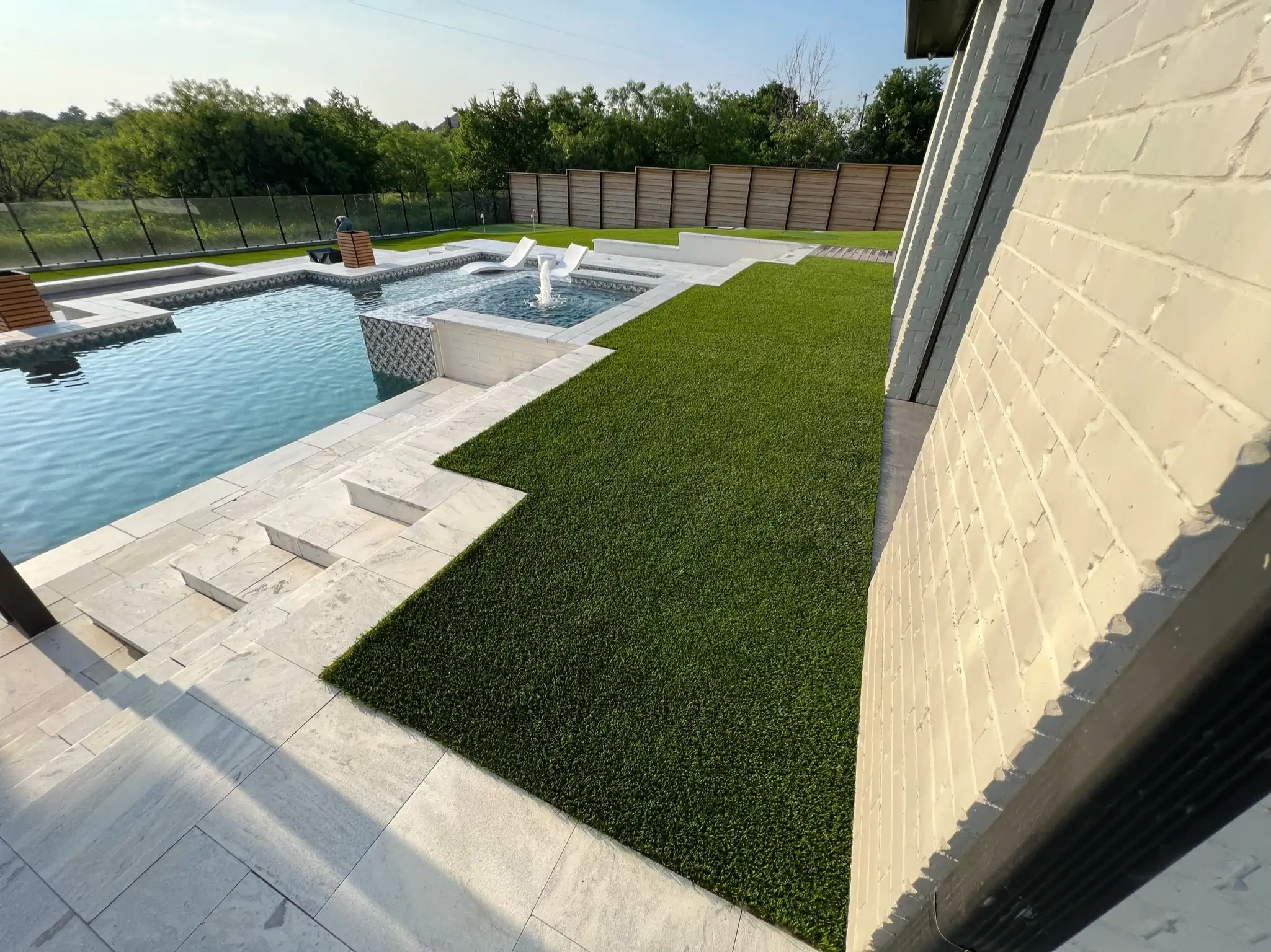 Elegant backyard with a rectangular swimming pool and marble coping, artificial grass lawn, and privacy fence in a suburban setting with the house's exterior wall in view.