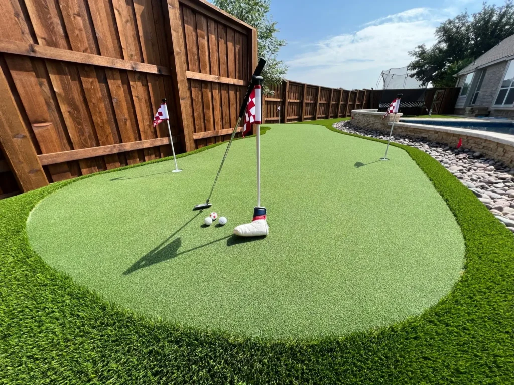 Close-up view of a backyard artificial turf putting green with two golf balls, a putter, and checkered flags, bordered by a decorative rock edge and a wooden fence under a clear sky.