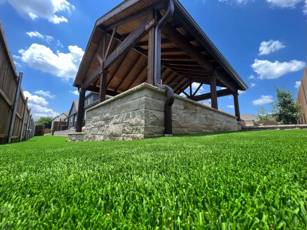 Low angle view of a backyard featuring vibrant artificial grass, with a stone foundation of a wooden gazebo against a clear blue sky, bordered by a wooden fence.