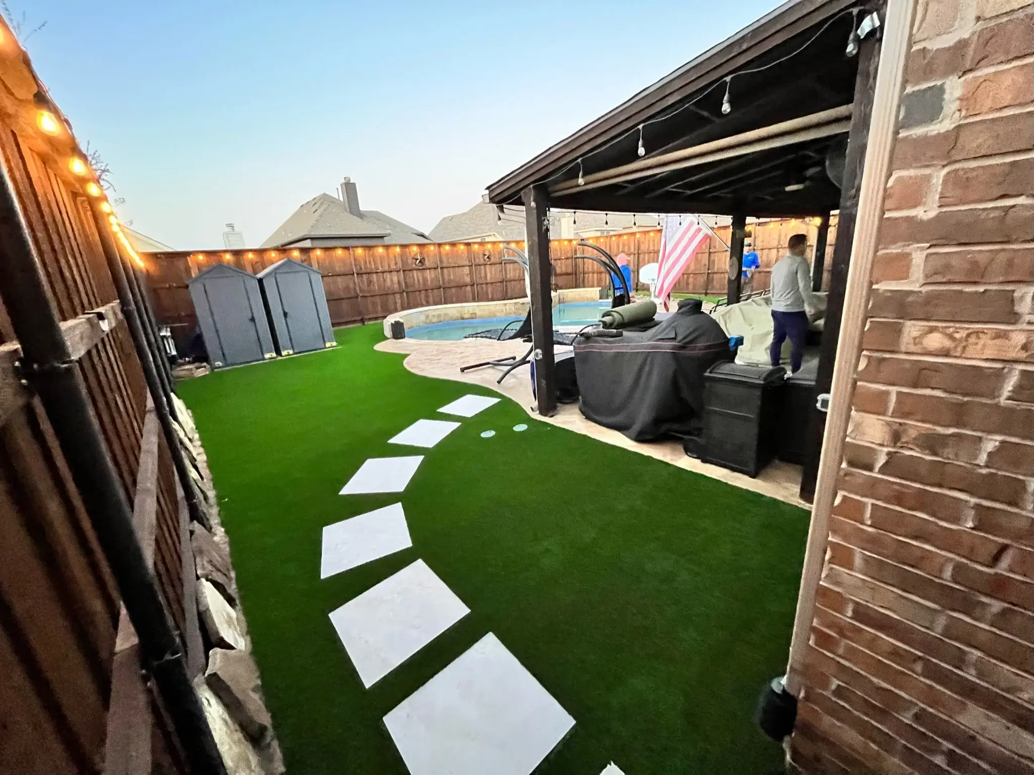 Backyard with artificial grass and pathway leading to a covered patio and pool area with ambient lighting at dusk.