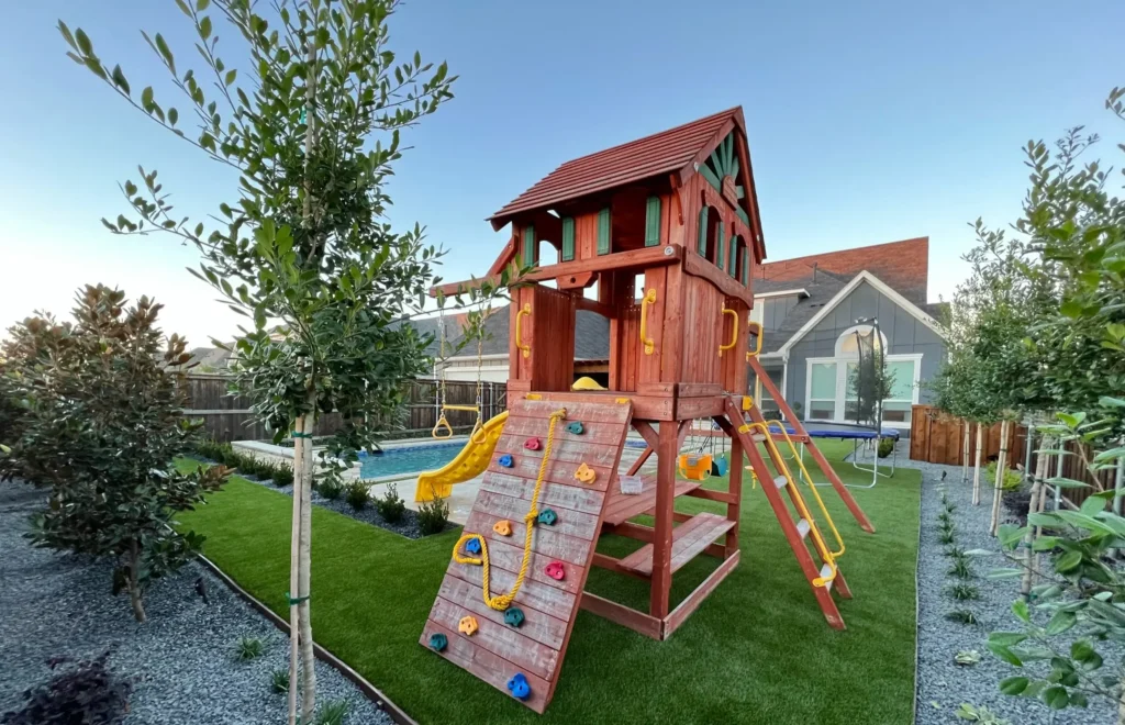 Backyard play area with a wooden playset featuring a slide, climbing wall, and swings on fresh green grass, complemented by landscaped garden beds and a backdrop of a residential pool and home.