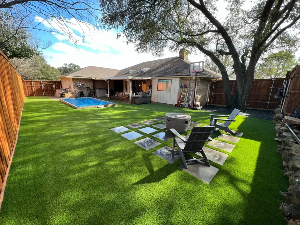 Expansive backyard with artificial grass and stepping stones leading to an inviting swimming pool, featuring outdoor lounging chairs and a fire pit, adjacent to a cozy home with a large oak tree.