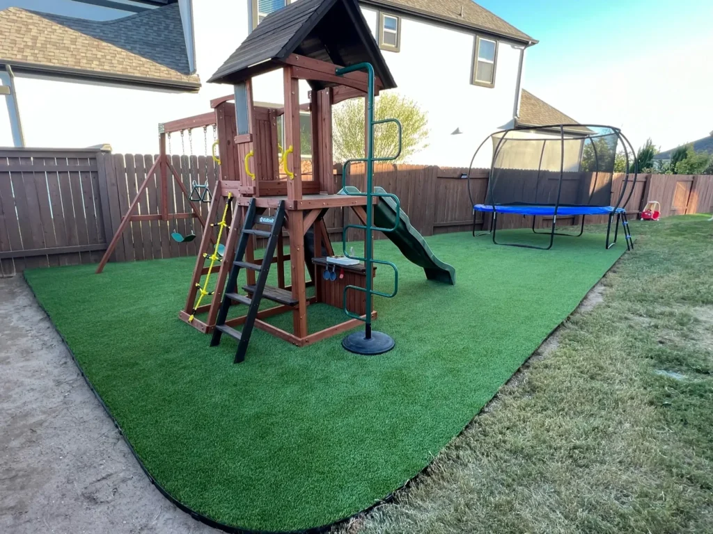Family backyard with a wooden playset including a slide, swings, and climbing steps, adjacent to a trampoline with safety net, on a well-maintained artificial grass patch bordered by a wooden fence.