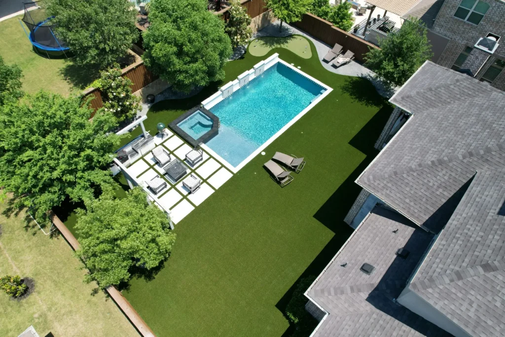 Aerial view of a residential backyard with a rectangular pool, integrated hot tub, and artificial grass surrounded by lush trees, alongside a modern house with gray shingles.