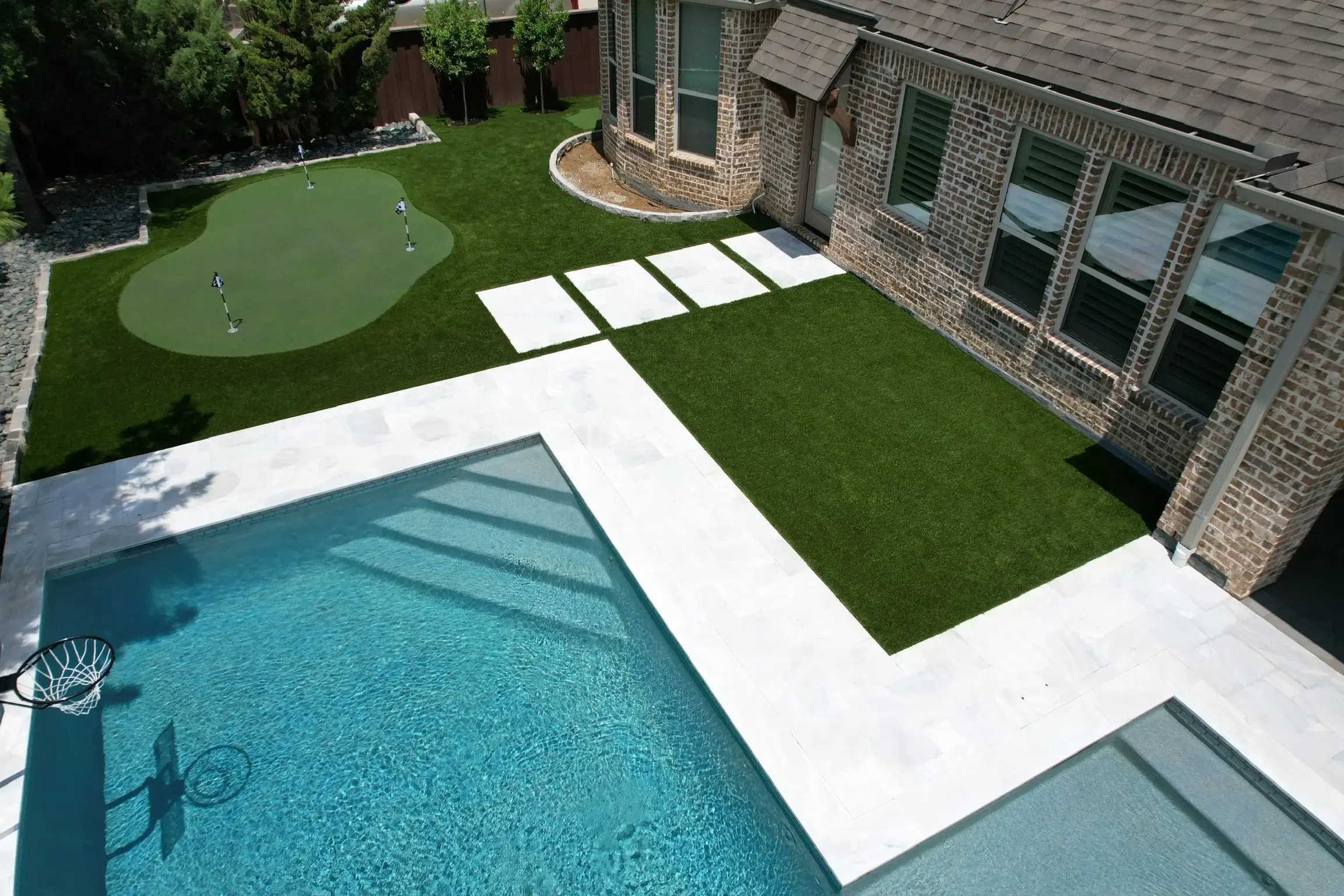 Luxury backyard with a clear blue swimming pool, basketball hoop, white stepping stones, artificial turf, and a small putting green, adjacent to a brick house with large windows.