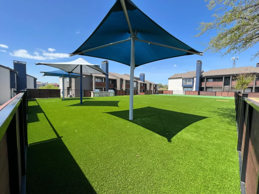 Community outdoor space with vibrant artificial turf and large blue sunshades, offering a recreational area in a residential neighborhood with modern townhouses in the background.