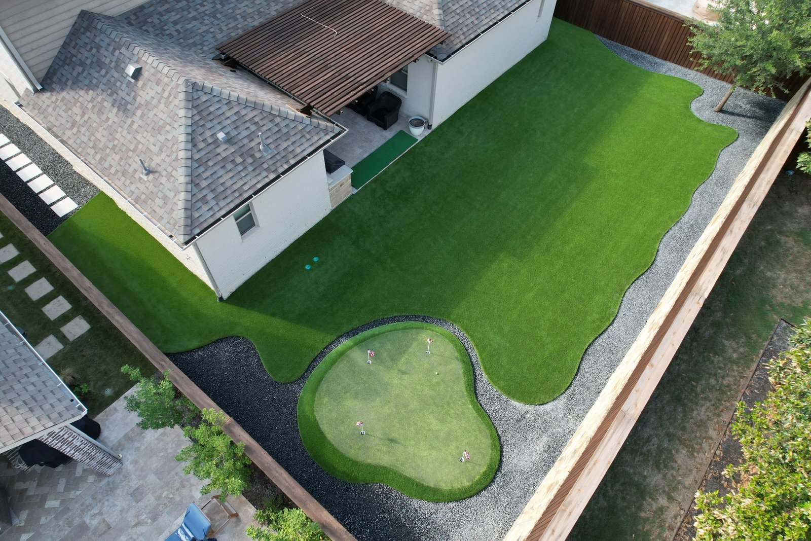 Aerial view of a residential backyard with custom-designed artificial grass, including a mini-golf green with a sand trap, surrounded by a wooden fence and adjacent to a home with a covered patio.