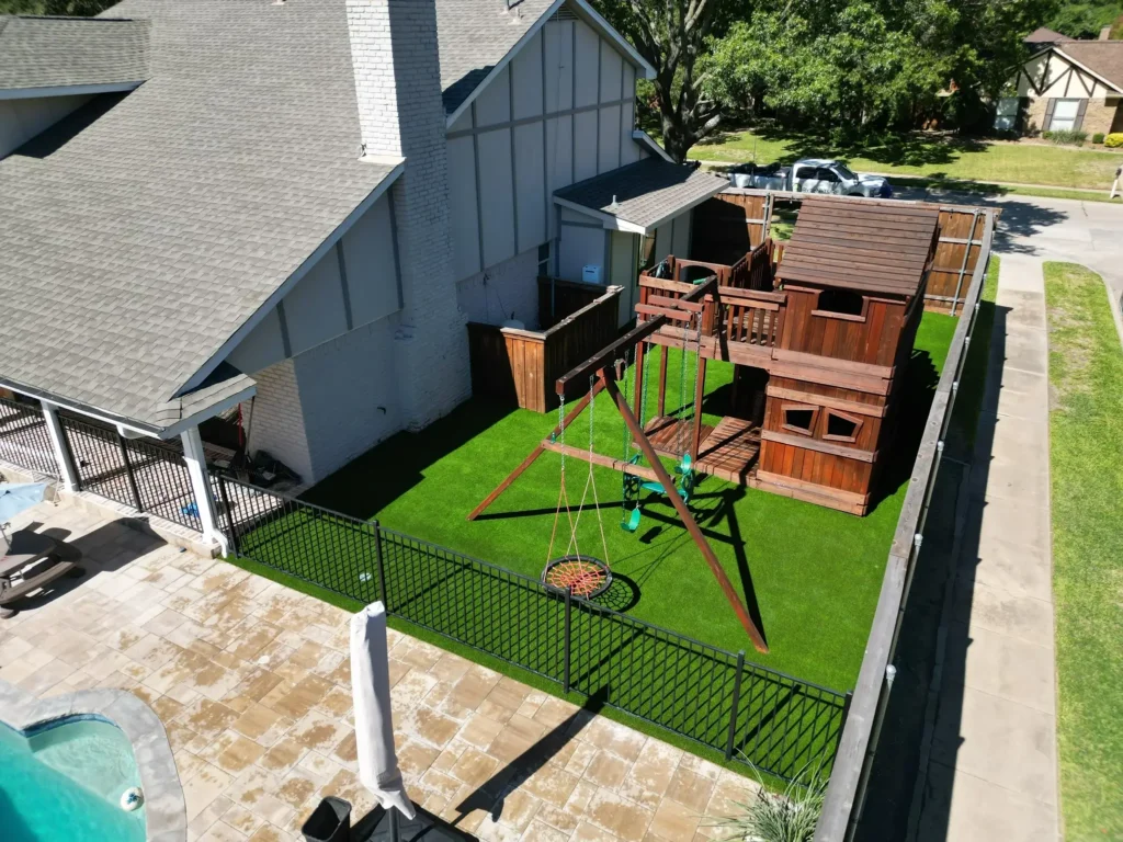 Aerial view of a residential backyard with a large wooden play structure with multiple levels and slides, a saucer swing, on vibrant artificial grass, adjacent to a patterned patio and a white-painted home.