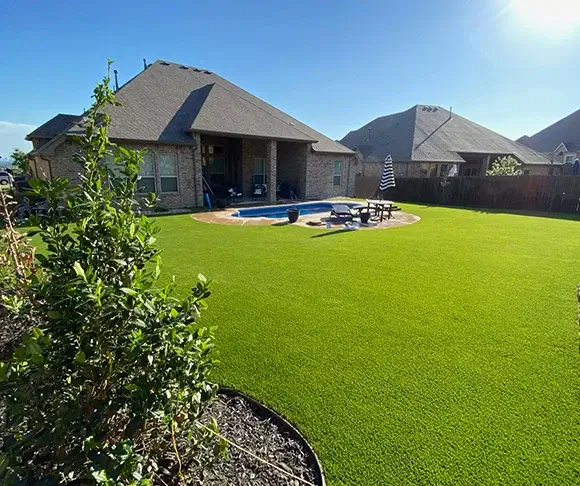 Spacious suburban backyard with vibrant green artificial turf, an in-ground swimming pool, and comfortable outdoor furniture, set against a backdrop of a modern single-story home with a dark shingle roof.