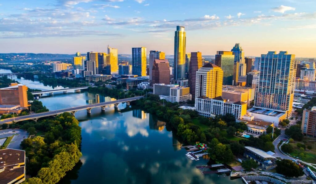 Aerial view of Austin, Texas skyline at sunset with Lady Bird Lake in the foreground, reflecting city buildings and connected by the Ann W. Richards Congress Avenue Bridge.