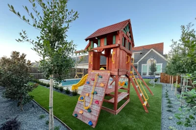Children's wooden playhouse with slide and climbing wall in a backyard garden, featuring greenery and a tree, during early evening.
